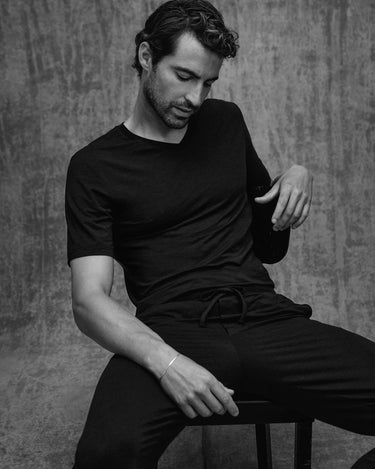 Man sitting on a stool wearing a black outfit against a textured gray background